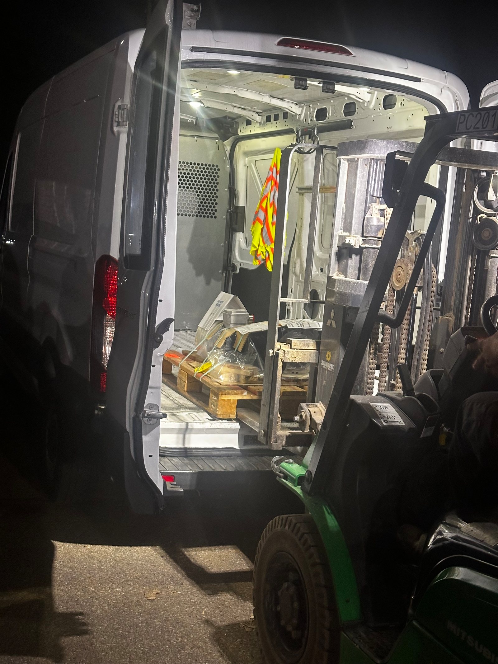 The client's own night frame in the yard: van doors open with the dome lights, a small palletized part on the floor, hi-vis